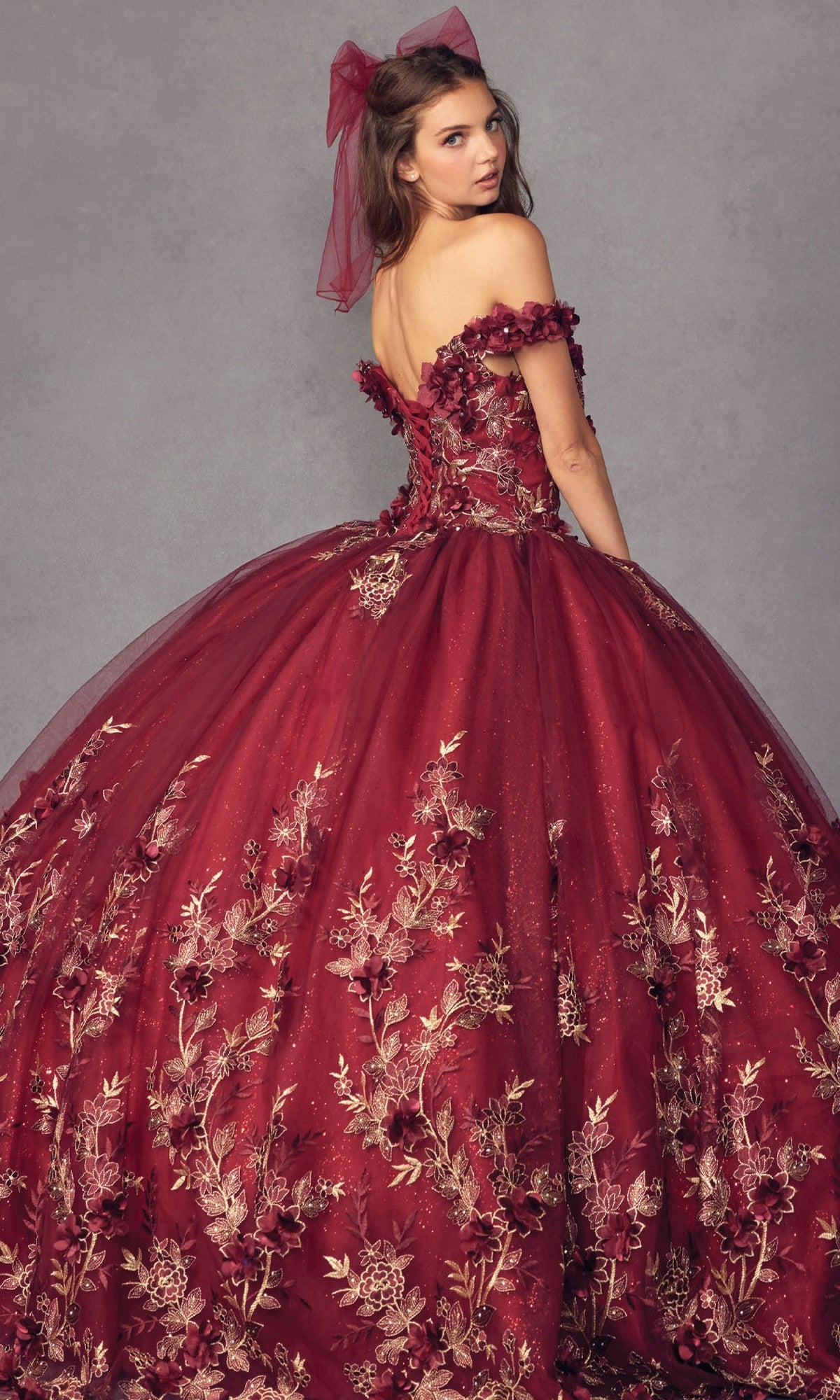 A young woman wears the Juliet 1447 Off-the-Shoulder Quinceañera Dress in red with gold beaded embroidery and a matching hair bow, looking over her shoulder against a gray background.